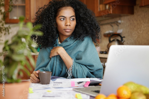 Worried beautiful African-American woman drinking coffee at kitchen table, planning budget, reviewing finances, calculating expenses and paying utility bills online, sitting in front of open laptop