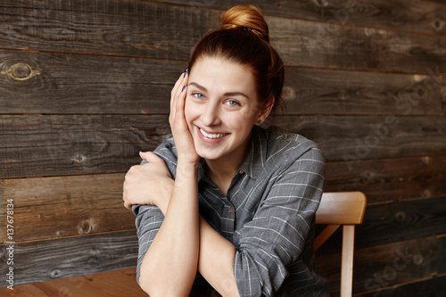 People and lifestyle concept. Portrait of good-looking girl with cute smile posing indoors against wooden wall background. Attractive young Caucasian woman with hair bun relaxing at cafe alone