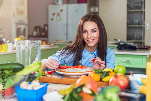 Pretty young woman smiling at camera while decorating a cake with sliced strawberry sitting in big kitchen