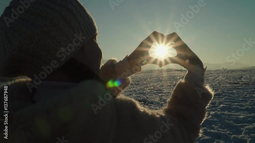 Child girl hands make heart around sun at sunset in mountains in sunny winter day
