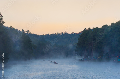 Beautiful view of People on Bamboo Boat with Fog in Lake and Pine Tree Background © komjomo