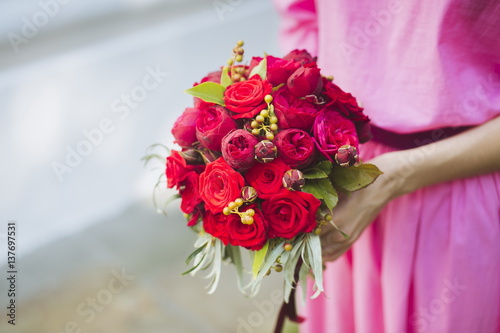 Happy bride in a simple pink wedding dress holding a beautiful bouquet of red roses, peonies and green leaves. Woman in a stylish dress celebrating summer day wedding, copy space, blurred background