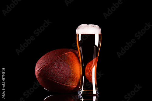 Beer glass with dark beer foam and American football ball on black background