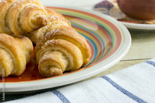 Cinnamon crescents in colored plate, a cup of coffee and cloth napkin on light wood background