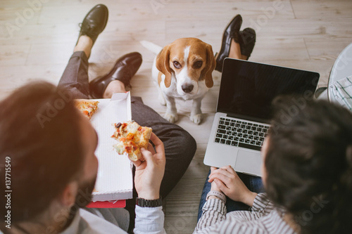 Young hipster couple home eating pizza with laptop and dog