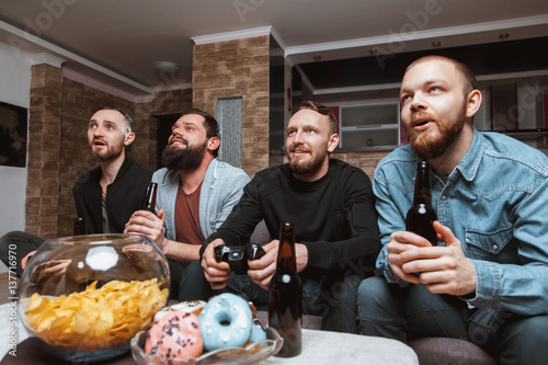 Men with a beard sitting on the couch at home talking and laughing with beer and joysticks in the hands of playing computer video games  one of them is eating a donut. 