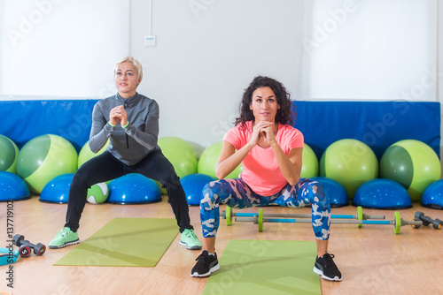 Two women in sportswear doing bodyweight squats while training indoors at sports hall