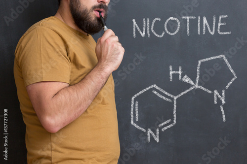 Nicotine molecule chemical structure on blackboard. Chemical structure of nicotine from cigarettes written on blackboard with man beside smoking electric cigarette photo