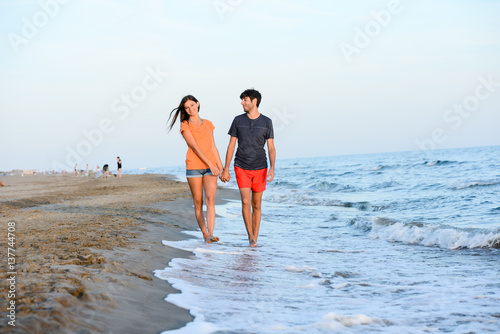 romantic young couple walking barefoot together in sand along the beach of mediterranean sea at sunset