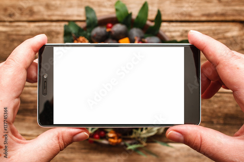 Food photography of plate with autumn fruits flat lay. Hands holding smartphone with blank screen above wooden background with figs dish. Instagram, networks, communication concept photo