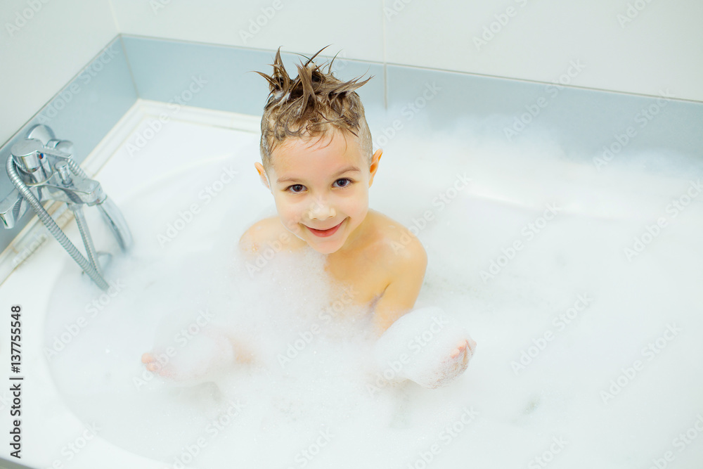 Happy young boy taking a bath in the foam. Smiling child washes his head with shampoo.
