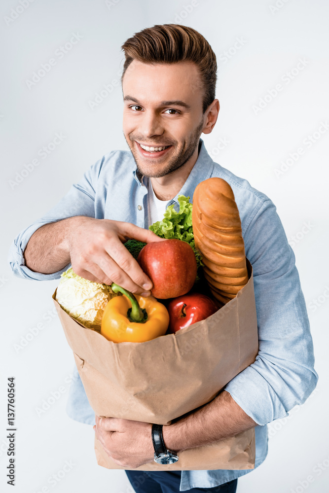Handsome young man holding grocery bag and smiling at camera Handsome young man holding grocery bag and smiling at camera
