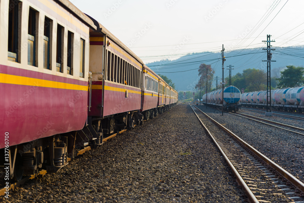 Fototapeta premium Passenger train of Thai Railways. Purple second class sitting car (in the left), fright tank cars and power poles (right) and a blue hill in the background. Train Ubon Ratchathani - Bangkok (Thailand)