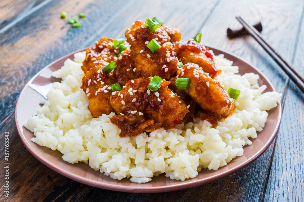 Crispy sesame chicken, chopped breast fillets, with a sticky sweet Asian sauce and white boiled rice on a plate on wooden table. Crispy sesame chicken, chopped breast fillets, with a sticky sweet Asian sauce and white boiled rice on a plate on wooden table.