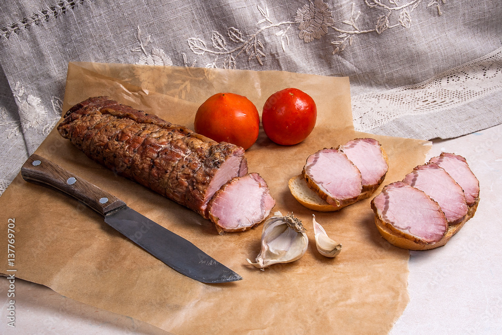 Slices smoked meat or ham and knife on brown packing paper. Tomatoes, garlic and small snacks of bread and meat.