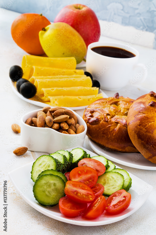 Breakfast on the table, salad from fresh tomatoes and cucumbers, almond nuts, bread, cheese, black olives, cup of coffee and fruits.