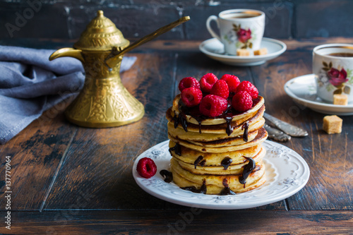 Stack of homemade pancakes with raspberries and chocolate, vintage white plate, Turkish coffee pot, forks, cups, brown sugar, wooden table.