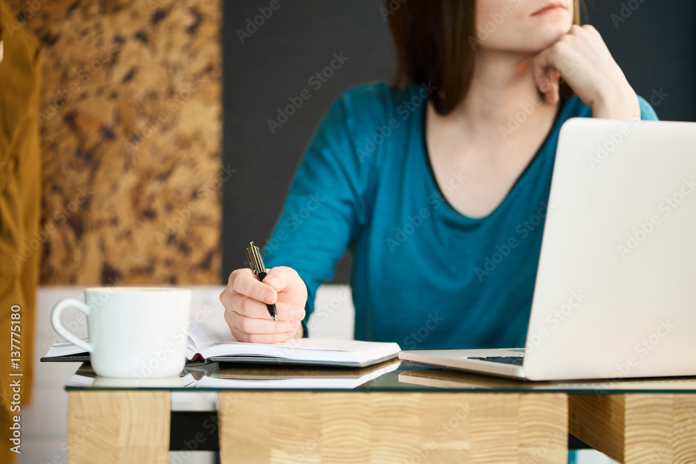 Young woman sits near the window at the cafe and working, she is making notes in her notepad, her laptop is open Young woman sits near the window at the cafe and working, she is making notes in her notepad, her laptop is open