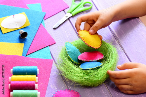 Small child does Easter decor. Child holds a felt egg in hand. Colorful felt Easter eggs and sisal nest, sewing tools and materials on a table. Simple and creative Easter decor idea