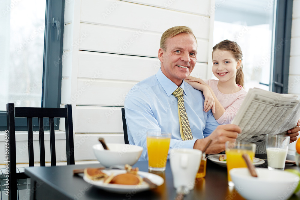 Businessman with newspaper and his daughter sitting by table in the kitchen Businessman with newspaper and his daughter sitting by table in the kitchen