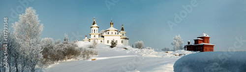 White orthodox church amidst deep snow and bare trees on the background of clear blue sky panorama