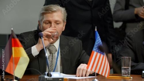 Representative of United States delegation get nervous at international summit of G20. Blonde middle aged politician man drinks mineral water, straighten his tie while prepare before speech. He photo