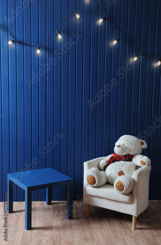 Big tedybear in scarf on white chair and blue table on a background of blue wall with retro garland of light bulbs. photo