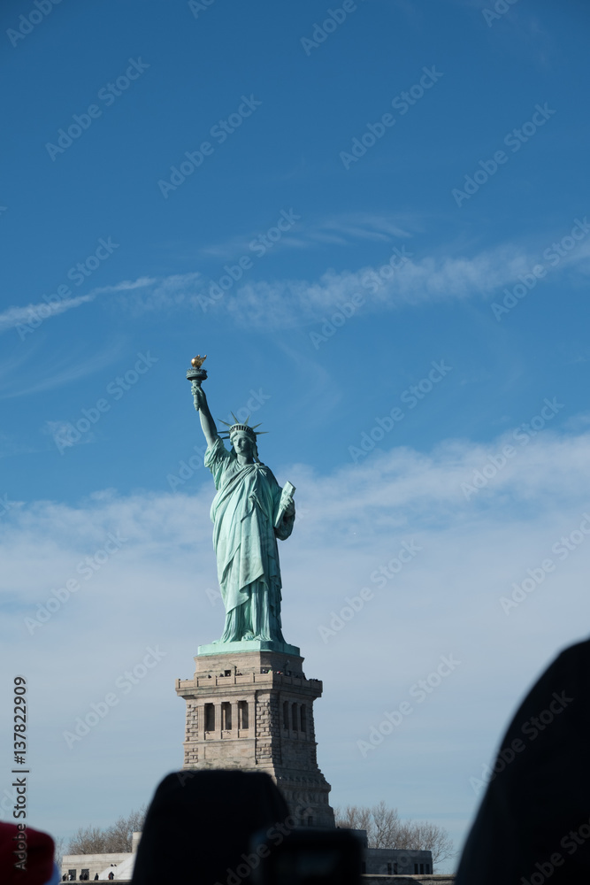 Obraz premium Looking at the Statue of Liberty from a crowded ferry, silhouettes of heads of people looking at Lady Liberty from a boat. Statue of Liberty on a bright sunny day with blue skies and a few clouds.