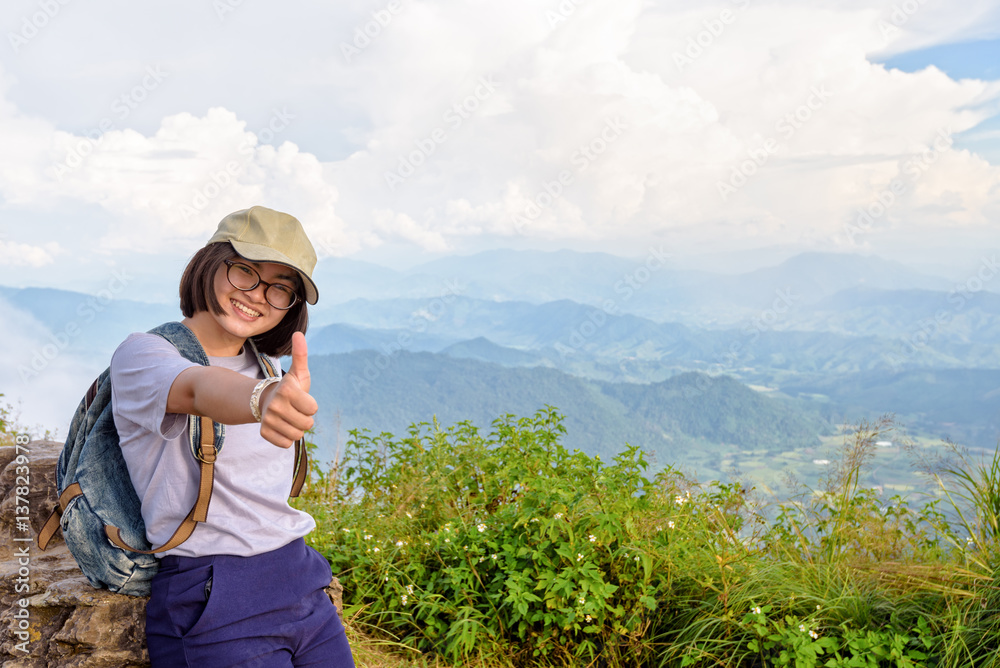Tourist teens girl hiker wear cap and glasses with backpack poses thumb up smiling happily on high mountain at scenic point of Phu Chi Fa Forest Park in Chiang Rai Province Thailand