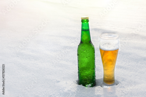 Mug and bottle of cold beer in the snow at sunset. Beautiful winter background. Outdoor recreation. Advertising of alcoholic beverage. Booze and holidays.