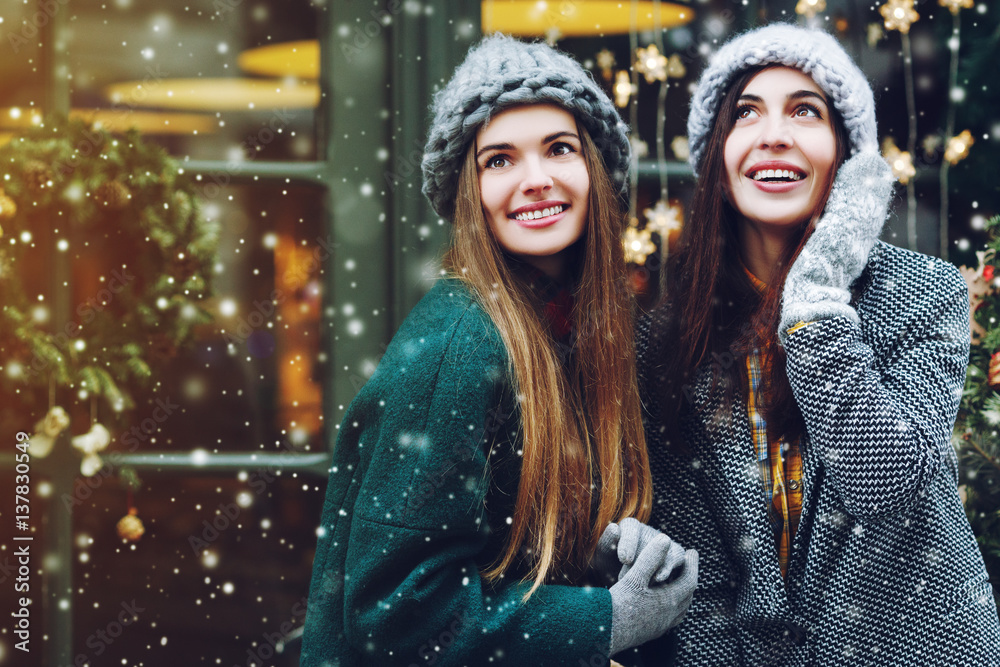 Outdoor waist up portrait of two young beautiful fashionable happy smiling girls posing on street. Models looking up, wearing stylish winter clothes. Snowfall, christmas background. Copy, empty space Outdoor waist up portrait of two young beautiful fashionable happy smiling girls posing on street. Models looking up, wearing stylish winter clothes. Snowfall, christmas background. Copy, empty space