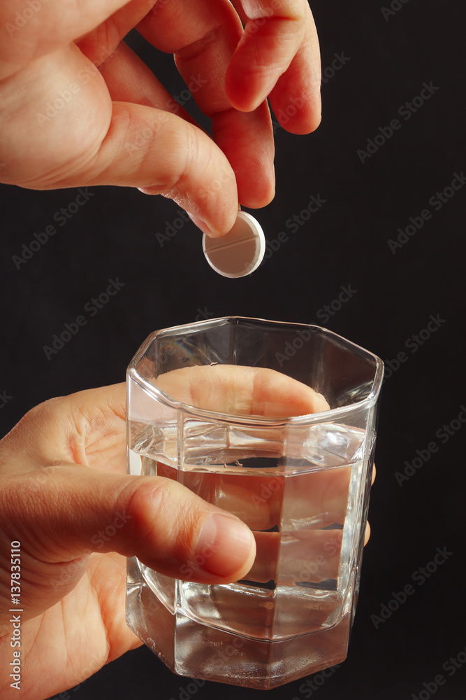 Hand throwing a soluble pill from headache in a glass of water on a dark background. Hand throwing a soluble pill from headache in a glass of water on a dark background.