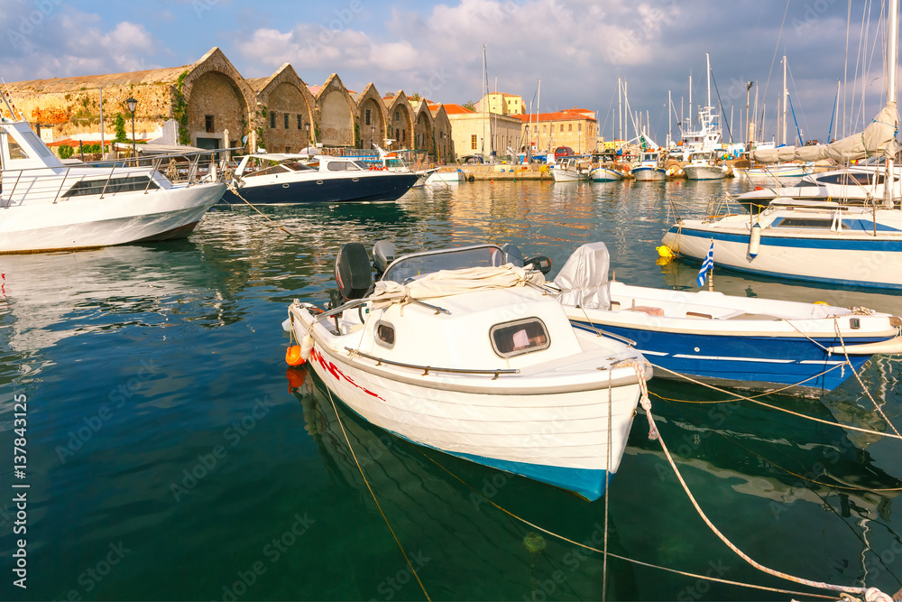Obraz premium Chania Arsenals, the Venetian shipyards, and fishing boats in old harbour of Chania in cloudy summer morning, Crete, Greece