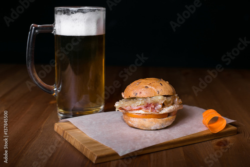 Fresh homemade burger with in old metal plate with fried potatoes in baking paper, served with glass of cold lager beer over old wooden table with dark background. Dark rustic style.