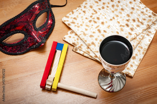 Judaism and religious jewish holiday with matzos (unleavened flatbread), cup of wine, wooden noisemaker or gragger (a traditional toy) for purim celebration and a red mask on wooden background