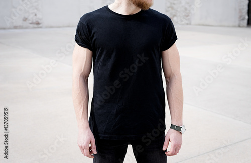 Young muscular bearded man wearing black tshirt and jeans posing in center of modern city. Empty concrete wall on the background. Hotizontal mockup, front view