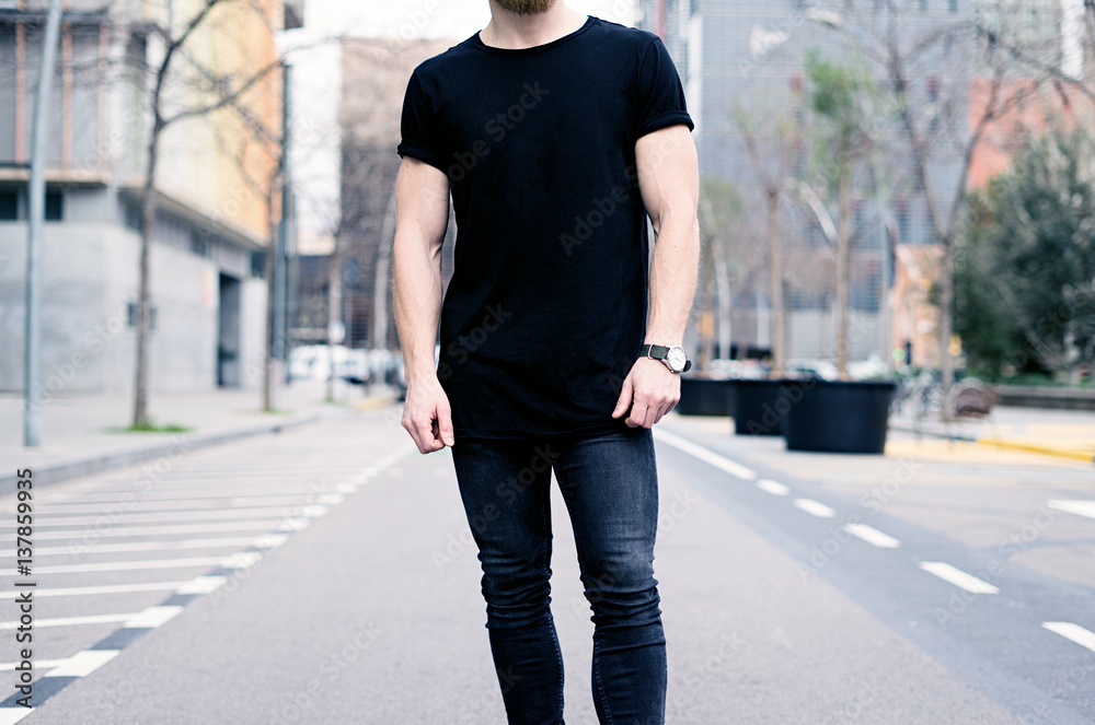 Young muscular man wearing black tshirt and jeans posing on the street of the modern city. Blurred background. Hotizontal mockup. Young muscular man wearing black tshirt and jeans posing on the street of the modern city. Blurred background. Hotizontal mockup.