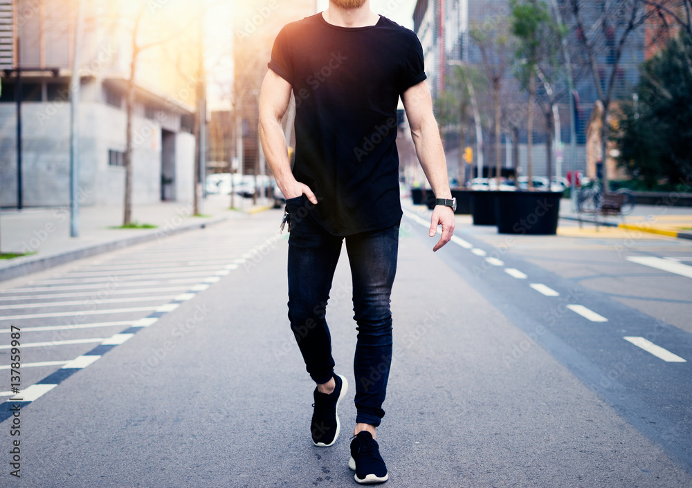 Young muscular man wearing black tshirt and jeans walking on the streets of the modern city. Blurred background. Hotizontal mockup.