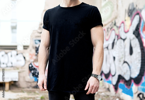 Young caucasian muscular man wearing black tshirt and jeans posing in modern district.Blurred graffiti on the background. Hotizontal mockup.