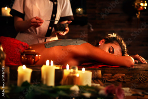 Mud mask of woman in spa salon. Back massage with clay full body . Girl on interior with candles foreground in oriental therapy room. Female lying on wooden spa bed. Beautician used cosmetic brush and