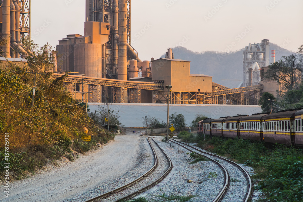 Cement factory in sunset lights. Cement plant in Saraburi province, Thailand. Rails can be seen as well as a passenger train passing by (Thai Railways)