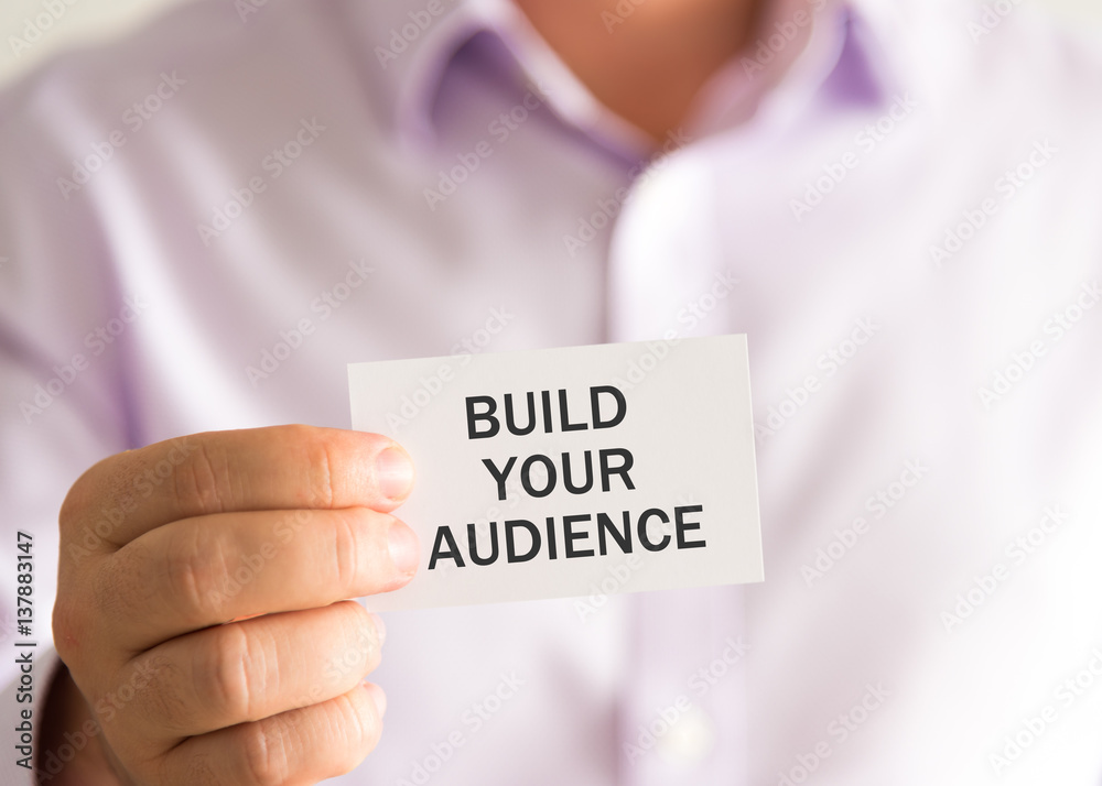 Businessman holding a card with text BUILD YOUR AUDIENCE Businessman holding a card with text BUILD YOUR AUDIENCE