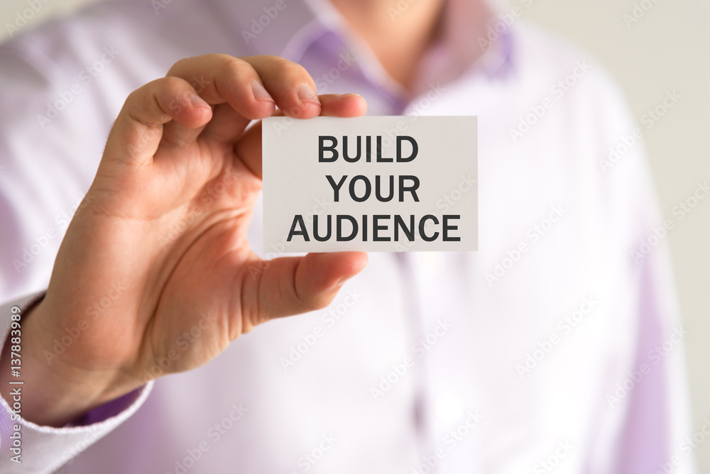 Businessman holding a card with text BUILD YOUR AUDIENCE Businessman holding a card with text BUILD YOUR AUDIENCE