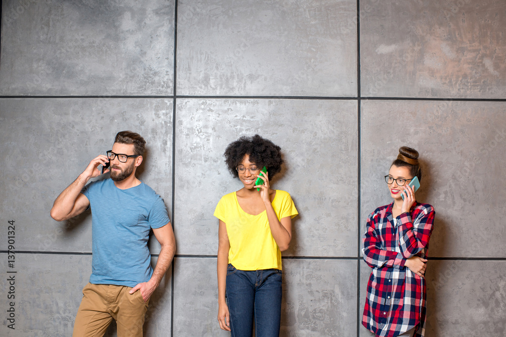 Fototapeta premium Multi ethnic coworkers dressed casually in colorful clothes talking with smart phones on the gray wall background