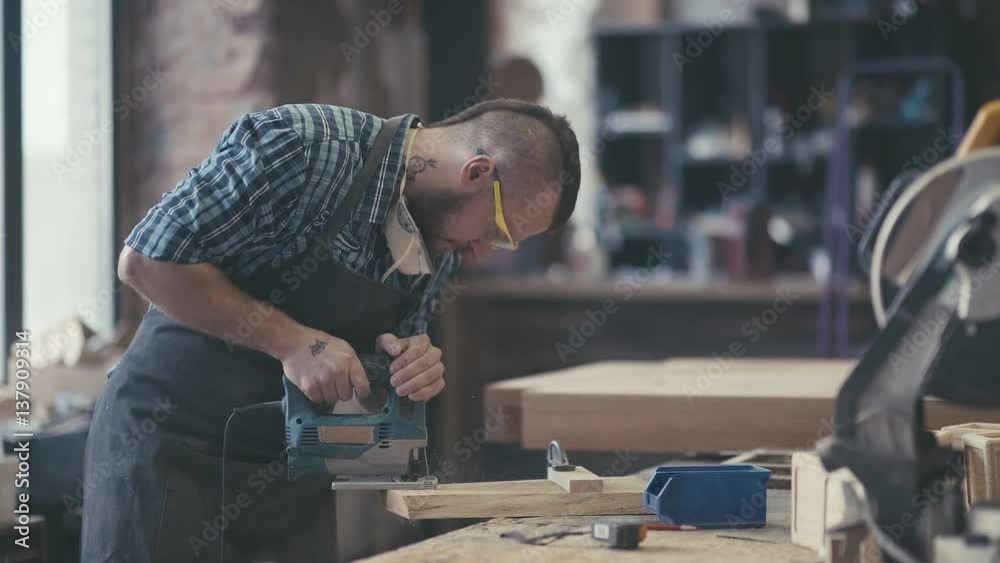 Carpenter with chisel in the hands on the workbench. Image of mature carpenter in the workshop. Carpenter cutting wooden board at his workshop. Woodman with tattoo. Carpenter with chisel in the hands on the workbench. Image of mature carpenter in the workshop. Carpenter cutting wooden board at his workshop. Woodman with tattoo.