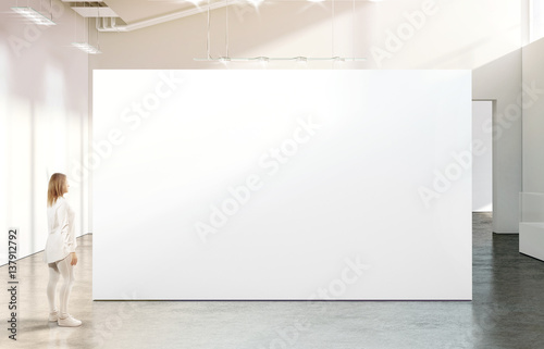 Woman walking near blank white wall mockup in modern gallery. Girl admires a clear big stand mock up in museum with contemporary art exhibitions. Large hall interior, banner exposition