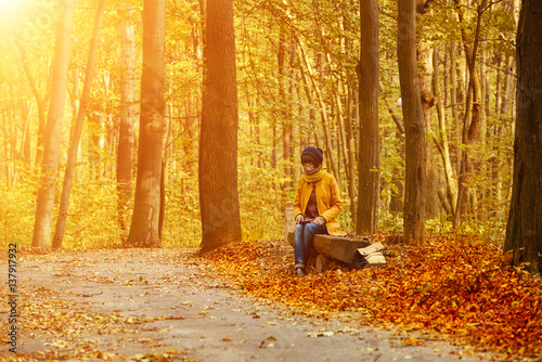 Young stylish hipster girl sitting on the bench in the autumn sunny park and reading electronic book