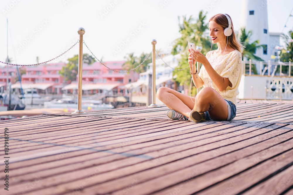 Enjoying the sound. Happy young woman with earphones is listening music while sitting on deck broadwalk in marine harbour.