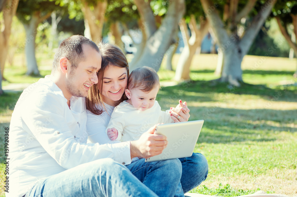 Young parents and little kid using table computer while sitting on grass Young parents and little kid using table computer while sitting on grass