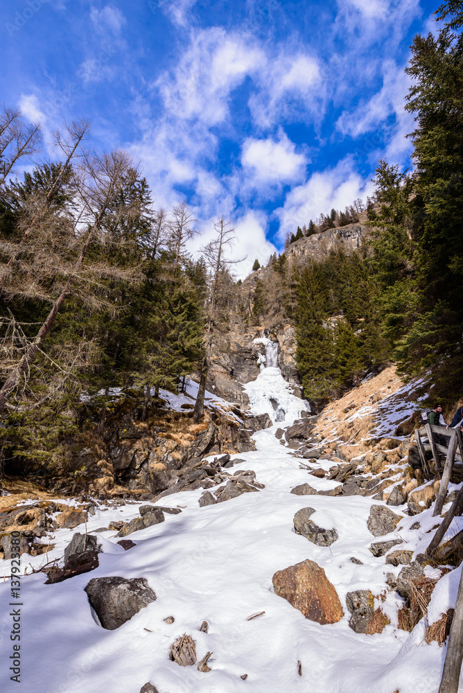 Saent waterfall, in Val di Rabbi, Trentino Alto Adige, covered by snow in a day with clear sky with few clouds. Trees are without snow. It a typical winter / spring mountain landscape/scenery